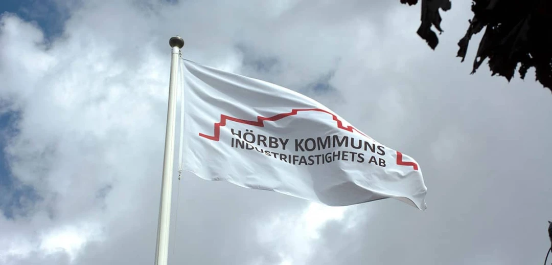 Vit flagga med Hörby Industrifastighets AB:s logotyp vajar på flaggstång